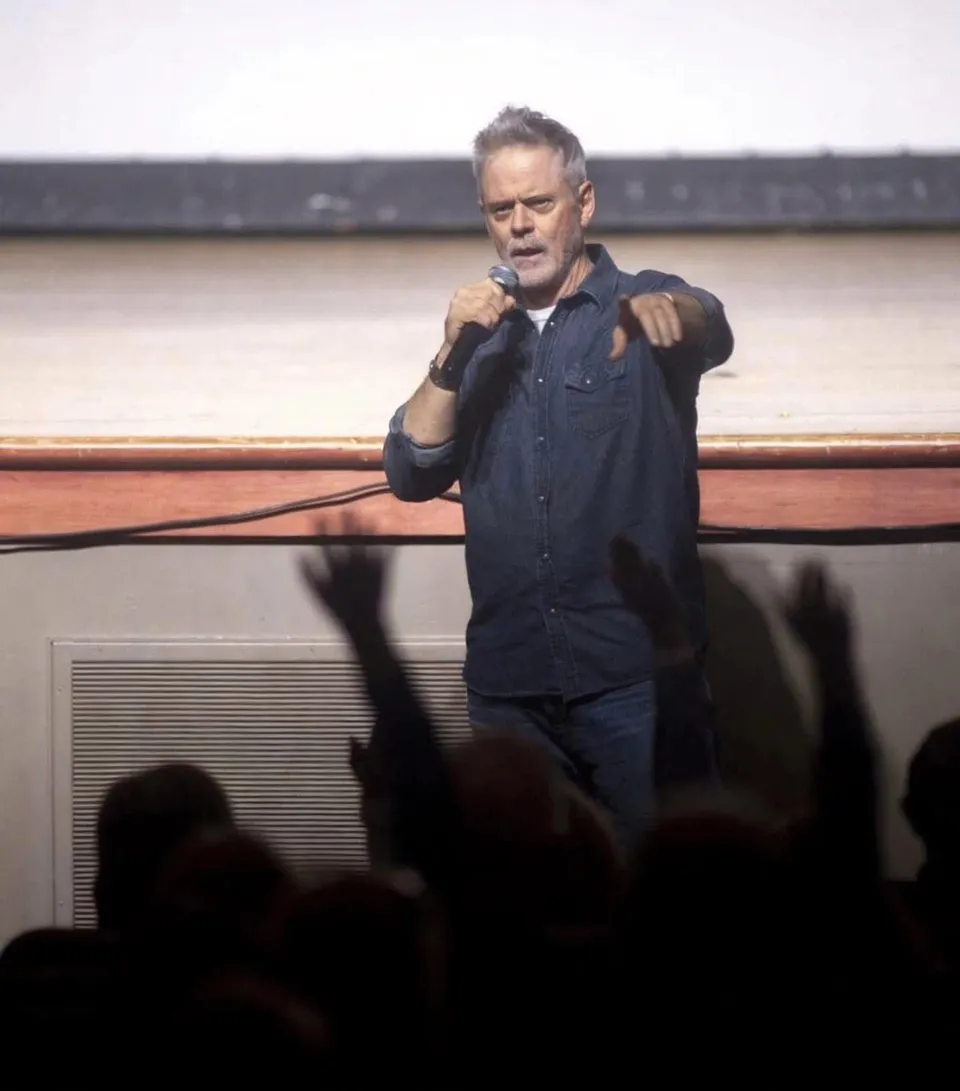 C. Thomas Howell speaking into a microphone on a theater stage as audience members raise their hands