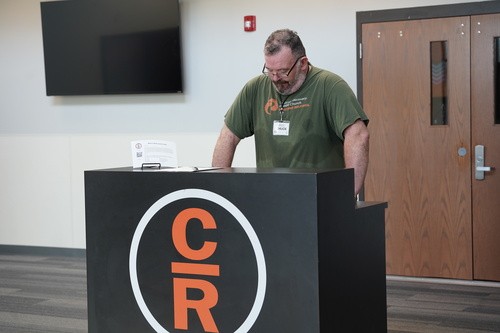 A man in a green T-shirt speaks from a podium marked with the orange Celebrate Recovery CR logo